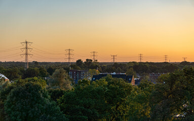 landscape at sunset, with several high-voltage power transmission towers stretching across the horizon. The foreground includes green trees and residential buildings, while the orange sky dominates
