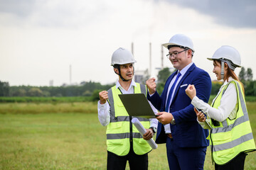 Three engineers, wearing helmets and reflective vests, celebrate a successful project discussion while reviewing plans on a laptop in an open field at an industrial site.