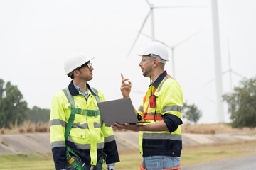 Male engineer using laptop computer at windmill field farm, wearing safety uniform and working and inspecting quality of wind turbines, repairing or maintenance wind turbines