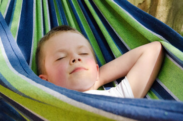 Cute little blond caucasian boy relaxing and having fun in multicolored hammock in backyard or outdoor playground. Summer active leisure for kids. Child swinging on hammock. Activities for children