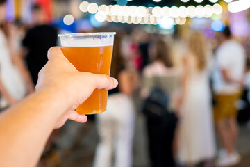 A hand holding a glass of beer at a social event, with a blurred background of people. Perfect for themes of social gatherings, leisure, nightlife, and celebrations