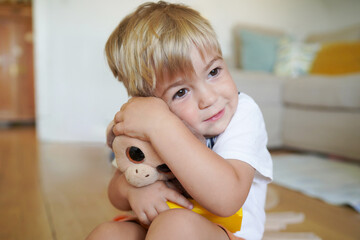 A cute young boy sitting on the floor, hugging his stuffed toy monkey with a gentle, loving expression in a cozy home setting.  © Татевик Багдасарян