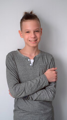 Portrait smiling teen boy on gray background in close pose with hands crossing on chest stands looking at camera. Optimistic emotional blond young guy with long hairs in ponytail feeling joy optimism.