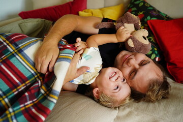 A happy father and young son cuddling on a couch under a blanket, with the boy holding a stuffed...