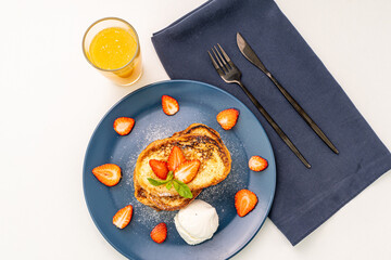 French toast decorated with strawberries and mint leaves with ice cream drizzled with caramel sauce and powdered sugar on a blue plate and a glass of orange juice on a white background, table