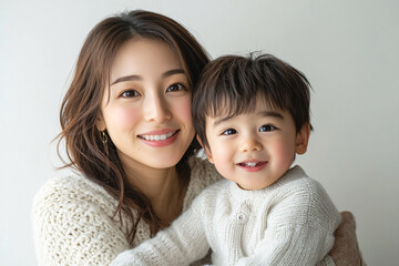 Young Japanese mother holding her cute young son, both looking into the camera, in front of a white studio background, showcasing a tender moment of family bonding and love