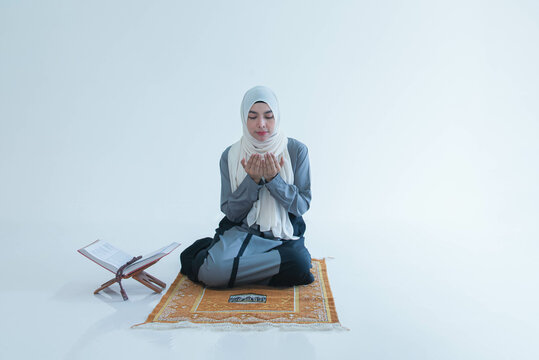 An Asian Muslim woman sits on a carpet in a white room and prays for a blessing that Muslims believe God grants to individuals and believers in Islam.