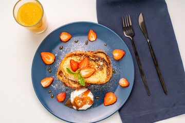 French toast decorated with strawberries and mint leaves with ice cream drizzled with caramel sauce and powdered sugar on a blue plate and a glass of orange juice on a white background, table