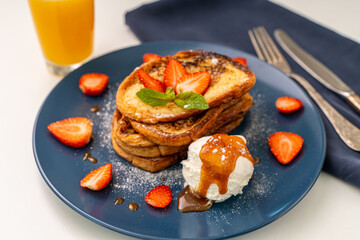 French toast decorated with strawberries and mint leaves with ice cream drizzled with caramel sauce and powdered sugar on a blue plate and a glass of orange juice on a white background, table