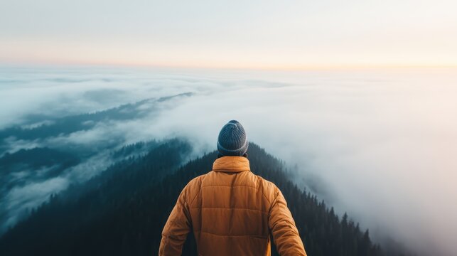 A person wearing an orange jacket overlooks a breathtaking mountain range with rolling clouds below, capturing the awe and solitude of being immersed in nature during a beautiful sunrise.