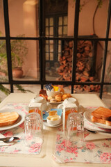 Pretty breakfast table setting in dining area by tall windows, in front of garden courtyard with firewood. Rustic cozy mood in hotel, hostel stay or guesthouse in Colonia del Sacramento, Uruguay