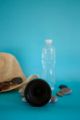 lens, straw hat, sunglasses, bottle of water, sunscreen spray, pebbles on a colored background