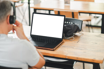 Man with headphones working on a laptop with a blank screen in a modern cafe. Wooden table and...