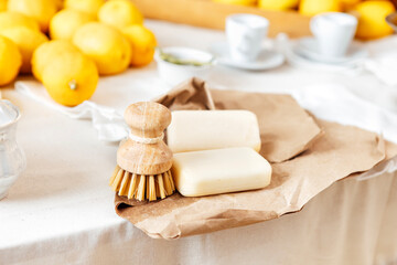 Close-up of two pieces of handmade soap and bamboo scrub brush for washing dishes on craft paper. Lemons and cups on table.
