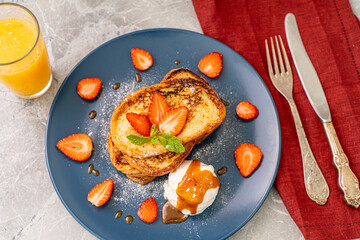 French toast with strawberries and mint, ice cream drizzled with caramel sauce on a blue plate with cutlery on a burgundy napkin orange juice on a gray background. Healthy breakfast concept.