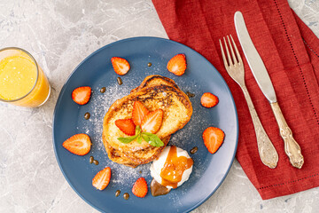 French toast with strawberries and mint, ice cream drizzled with caramel sauce on a blue plate with cutlery on a burgundy napkin orange juice on a gray background. Healthy breakfast concept.