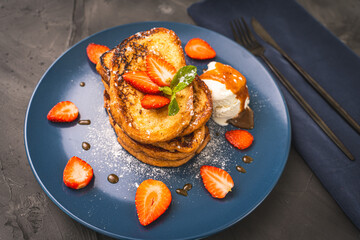 French toast with strawberries and mint, ice cream drizzled with caramel sauce on a blue plate with cutlery on a napkin on a gray background. Healthy breakfast concept.