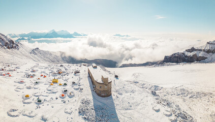 Aerial panoramic view Bethlemi Hut or meteo station and base camp acclimatization zone with tents for climbing Mount Kazbek. Kazbegi municipality. Snowy Caucasus mountain panorama in sunny day