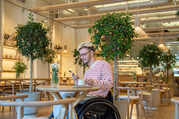 Thoughtful man in wheelchair drinking coffee at cafe table, using smartphone. Guy take break from...