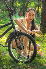 Fototapeta premium Beautiful Asian girl pumps a bicycle wheel with a pump in a park or forest at sunset. Bicycle repair. Healthy lifestyle.. Close-up