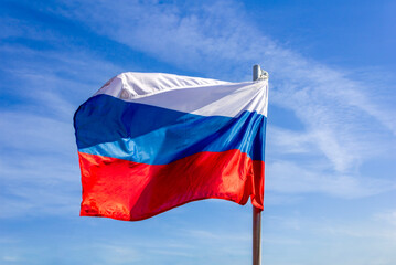 A clear daytime view of the Russian national flag waving in the wind against a blue sky.