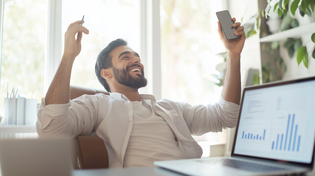 proud middle-aged Hispanic man, leaning back in his chair with an elated look, holding his smartphone up in one hand while a laptop displays business graphs on his desk, capturing