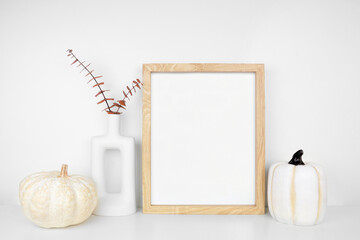 Mock up portrait wooden frame with fall branches and pumpkin decor on a white shelf against a white...