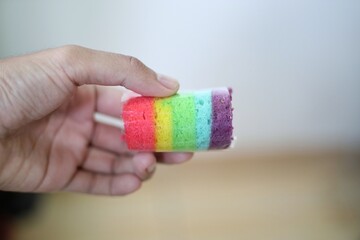 A hand is holding a colorful rainbow roll cake. The cake is wrapped in clear plastic and has vibrant layers of red, orange, yellow, green, blue, and purple.