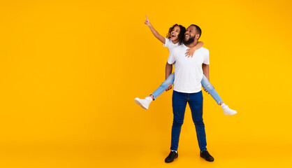 Great Offer. Full Body Length Of Cheerful African American Man Giving Piggy Back Ride For His Daughter, Little Girl Looking Aside And Pointing Fingers Up At Free Copy Space On Yellow Background © Prostock-studio