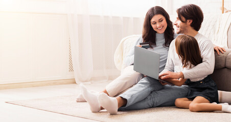 Obraz premium Booking vacation together. Happy young man, woman and their little daughter sitting on floor with laptop and credit card, making online reservation