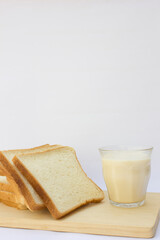 A clear glass of milk sits on the right side of a wooden cutting board, accompanied by three slices of white bread stacked to its left.