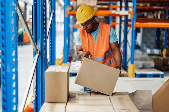 African american male warehouse worker handling box with care in stockroom aisle