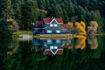 A distant photo of a wooden lake house on the water in the forest, taken from the opposite angle. The view of the house reflected in the water. GÃ¶lcÃ¼k Nature Park Bolu, Turkey.
