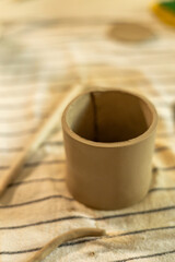 Close-up of a freshly crafted clay cup on a striped fabric surface. The unfinished ceramic piece highlights the hand-built pottery process, surrounded by crafting tools and clay remnants.