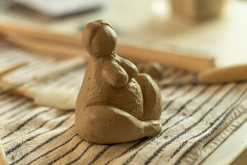 Close-up of a handmade clay sculpture depicting a seated human figure. The unfinished pottery piece...