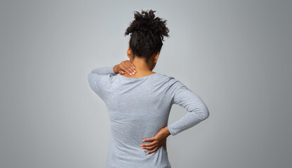African woman with neck and back pain, rubbing her painful body over grey background, back view