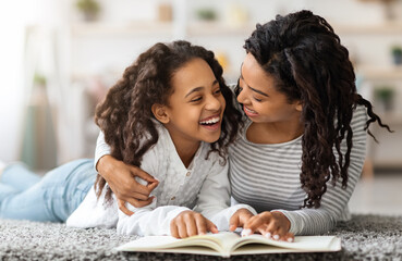 Fototapeta premium Joyful black girl reading book with her mom, laying together on floor and laughing, home interior. Happy african american mother and daughter reading books and embracing, closeup