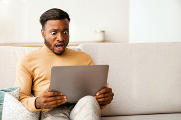 Shocked Black Guy Looking At Laptop Computer Working Sitting On Couch At Home. Selective Focus