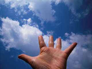 A close-up of a human hand reaching towards a bright blue sky filled with scattered white clouds. The image symbolizes aspiration, freedom, and connection with nature, shot from a low-angle perspectiv