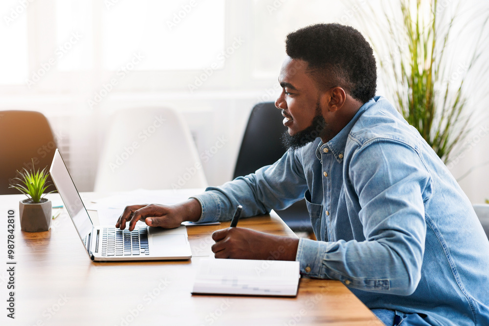 Poster african worker taking notes while using laptop in office, side view with free space