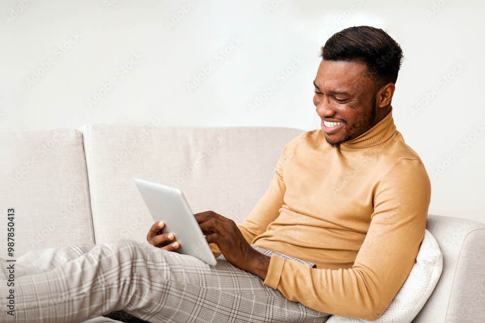 Fototapete happy african american guy using tablet computer browsing internet sitting on couch at home. selecti