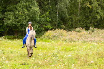 Fototapeta premium Lady in blue dress riding on Icelandic horse in tall hay. Rider is wearing helmet.