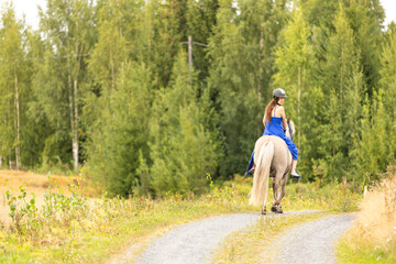 Lady with blue dress riding on Icelandic horse during sunset in Finland. Rider is wearing helmet.