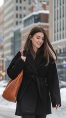 Fototapeta premium Woman in a black coat with a brown bag walking on a snowy city street, with buildings in the background. Concept of winter urban lifestyle