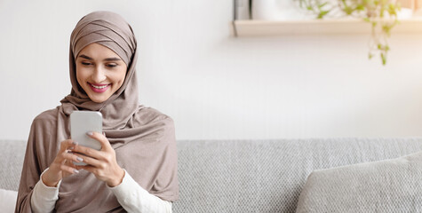 Portrait of millennial muslim girl in hijab using smartphone at home, messaging or browsing social...