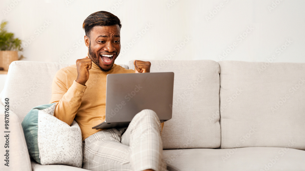 Poster yes. excited afro man using laptop computer shaking fists celebrating success sitting on sofa indoor