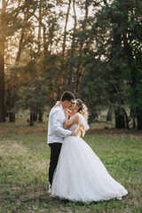 Fashionable groom and beautiful smiling bride curly blonde in white long dress hugging kissing outdoors in forest in nature evening at sunset. Wedding photography, portrait of happy newlyweds.