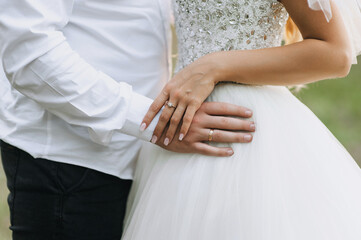 Groom in white shirt with bracelet and bride in dress with gold ring on finger standing outdoors holding hands. Close-up wedding photo of happy newlyweds.