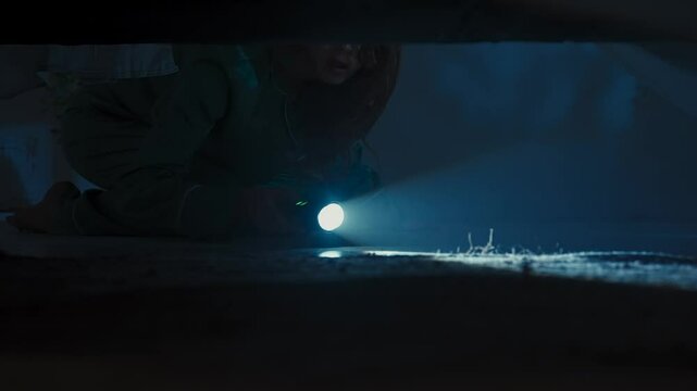 Woman on the floor in dark room checking space under bed with flashlight, looking around for monster, view from underneath bed. Halloween horror concept.