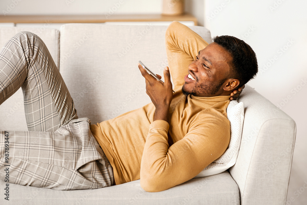 Poster cheerful african american guy using smartphone voice assistant or loudspeaker resting on couch at ho